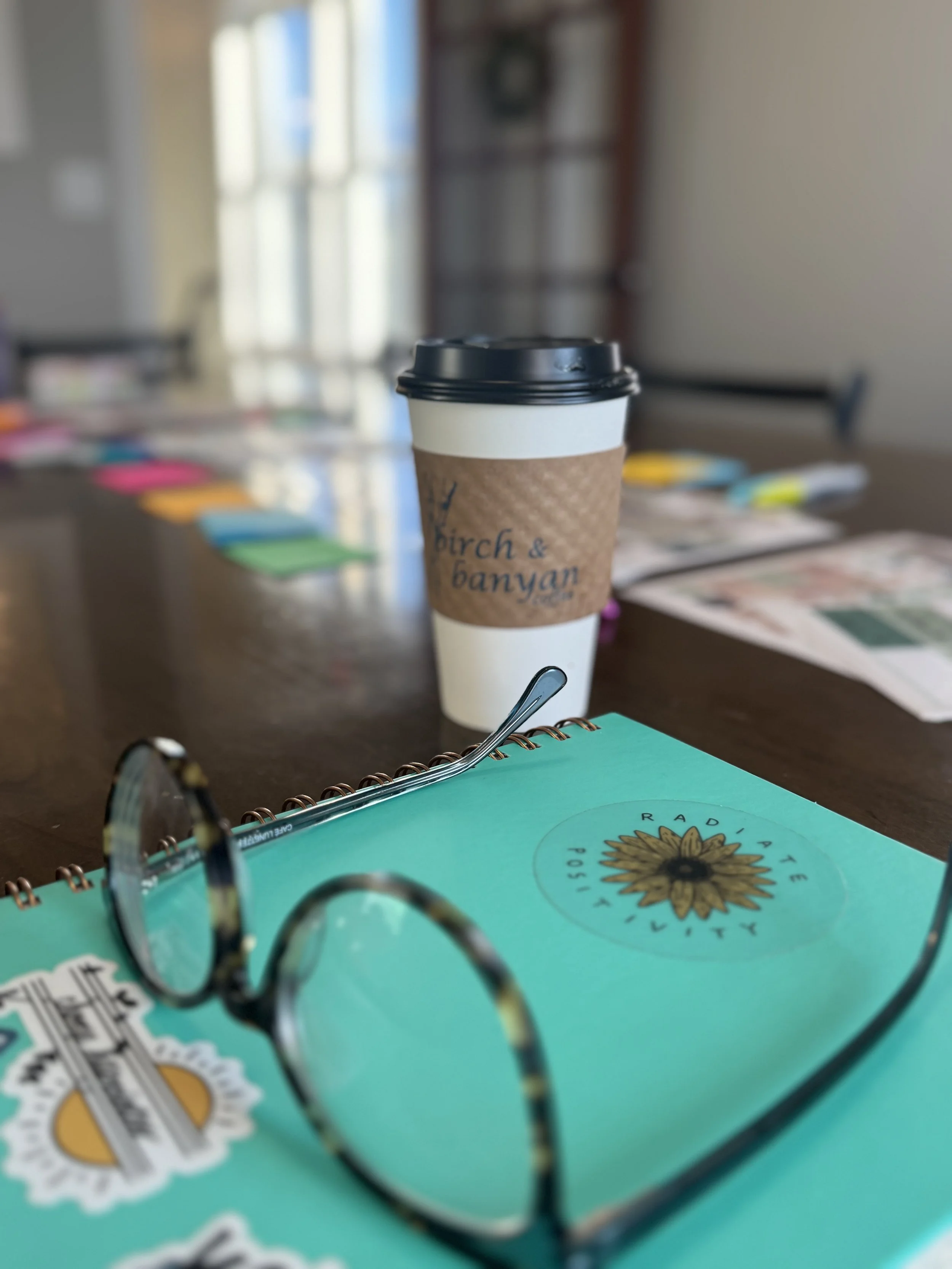 A disposable coffee cup with a black lid and a brown sleeve, placed on a table with a turquoise notebook and a pair of glasses nearby.