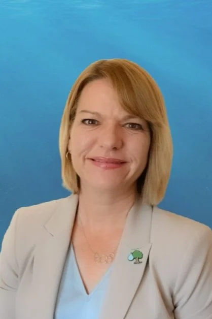 A woman with blonde hair wearing a light blazer and a necklace with a tree pendant, standing against a blue background.