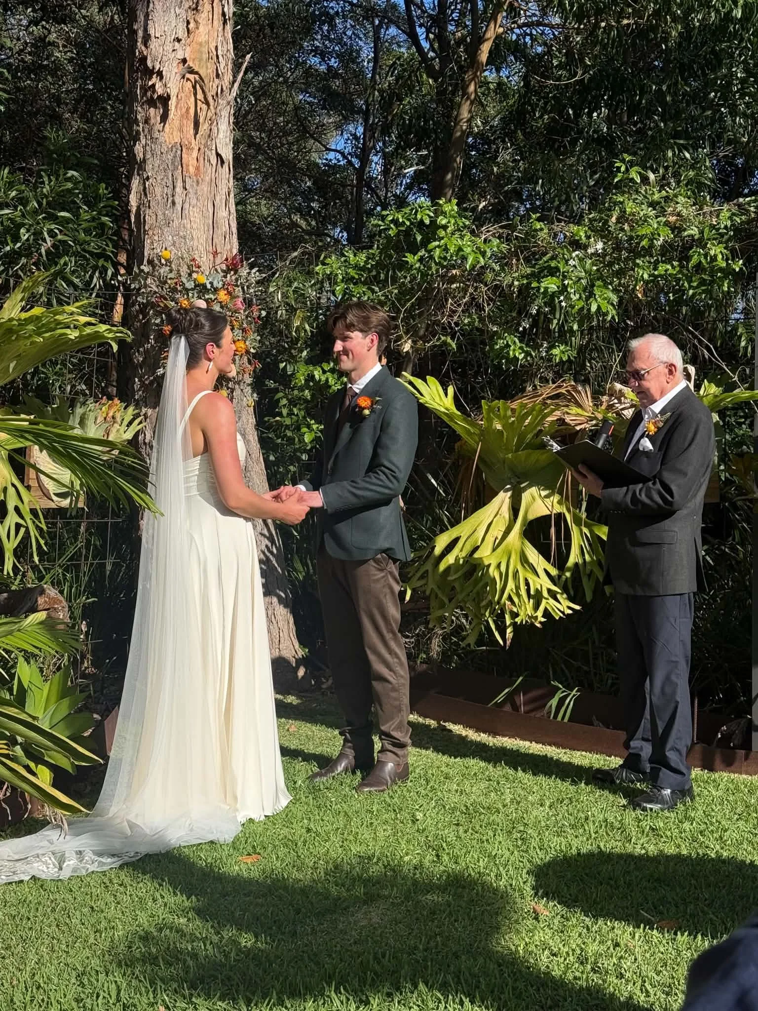 A wedding ceremony outdoors with a bride and groom holding hands and facing each other, a person officiating the ceremony, and a large tree and lush greenery in the background.