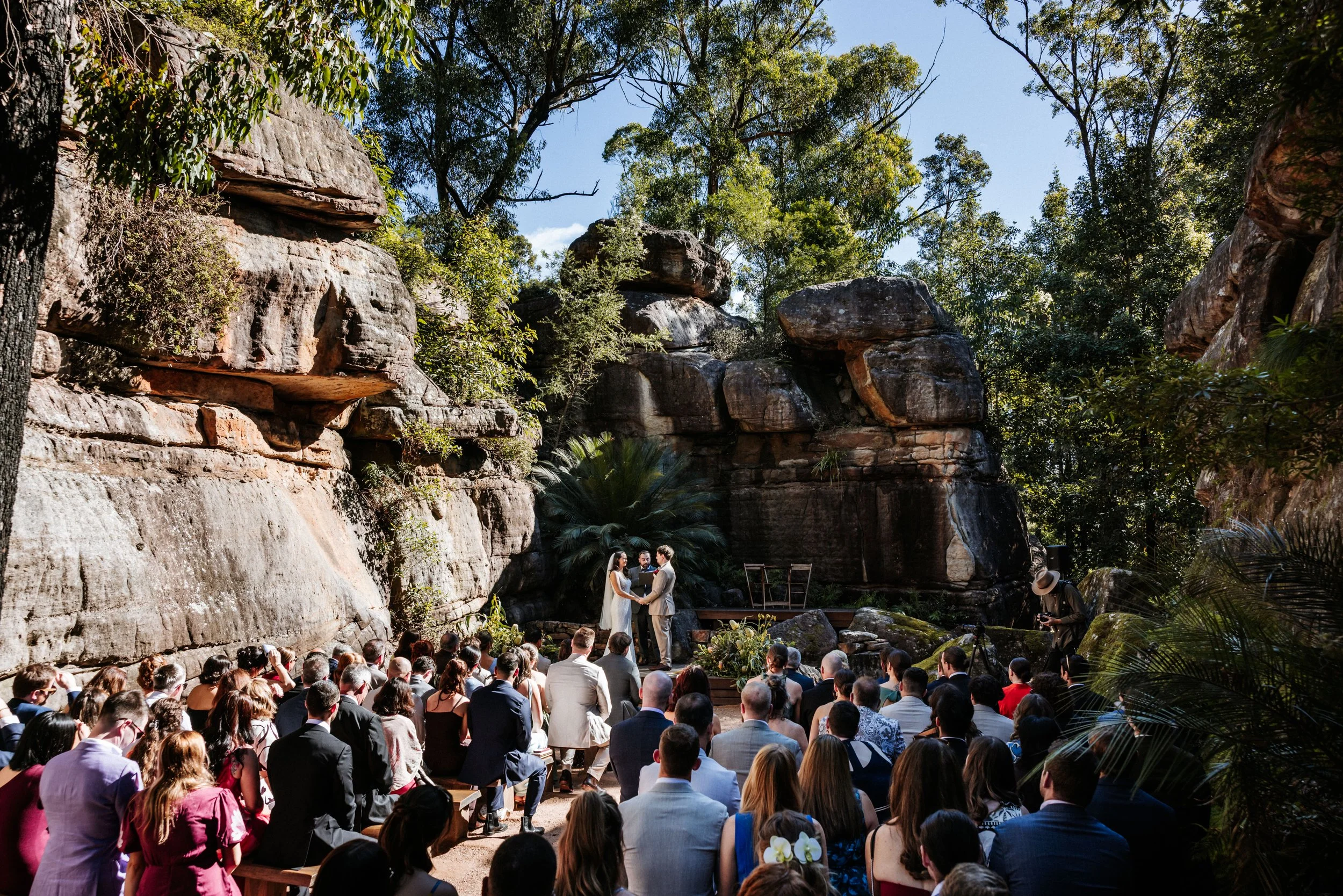 A wedding ceremony taking place outdoors beneath large rock formations and trees, with a large crowd of guests watching a couple exchange vows.
