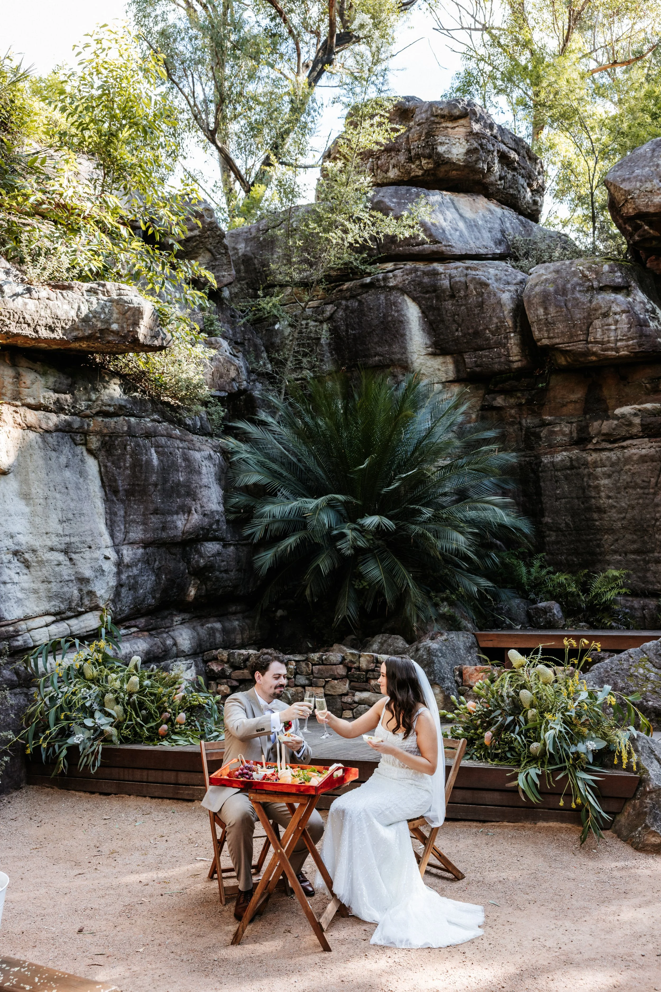 A bride and groom sitting outdoors at a wedding reception, raising glasses for a toast, surrounded by lush greenery and large rocks.