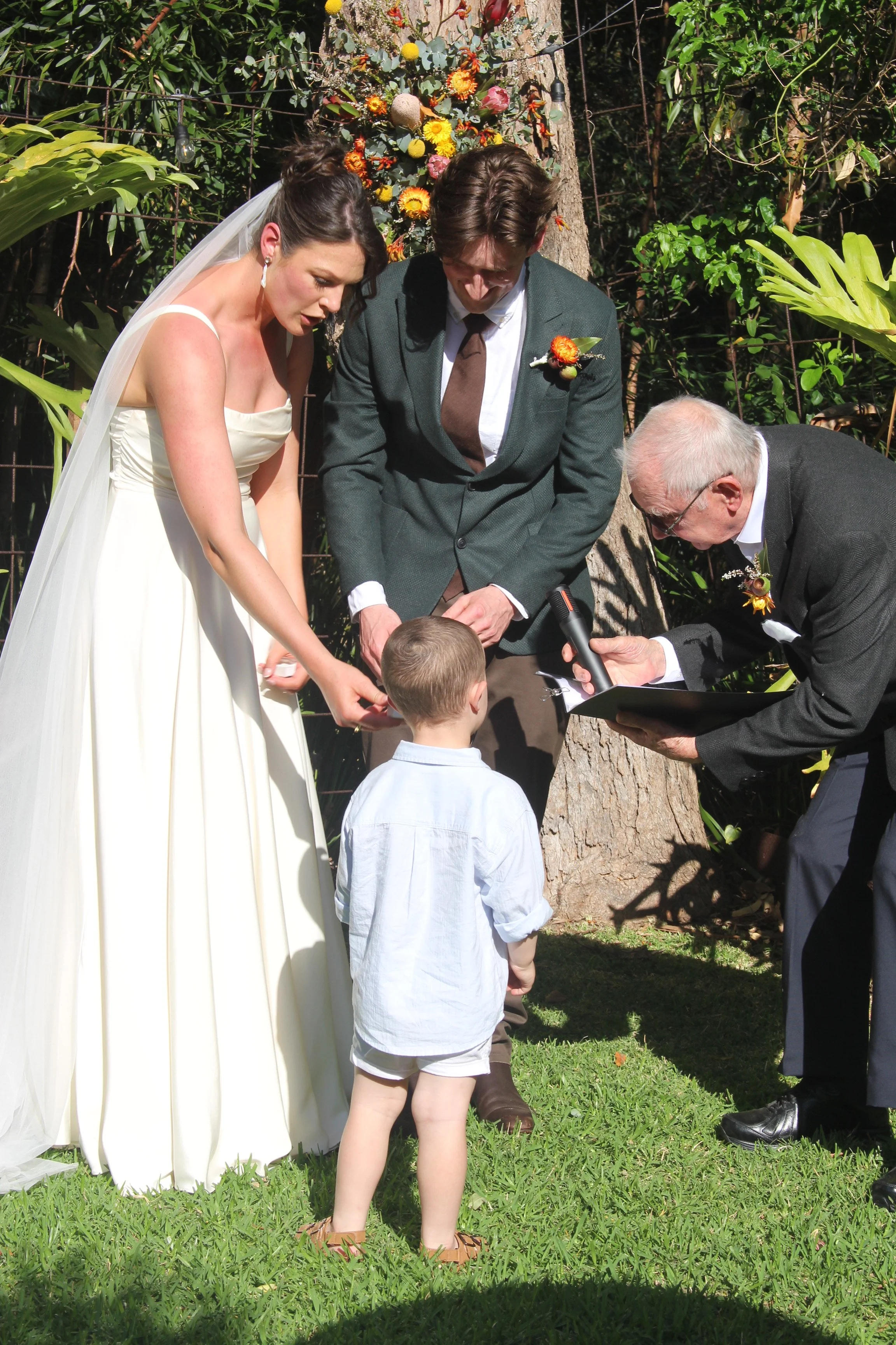 A wedding ceremony outdoors with a bride in a white dress and a veil, a groom in a grey suit, and an officiant in a black suit. A young boy stands in front of them, participating in the ceremony. Colorful flowers are arranged on a backdrop behind them, and there are trees and greenery surrounding the scene.