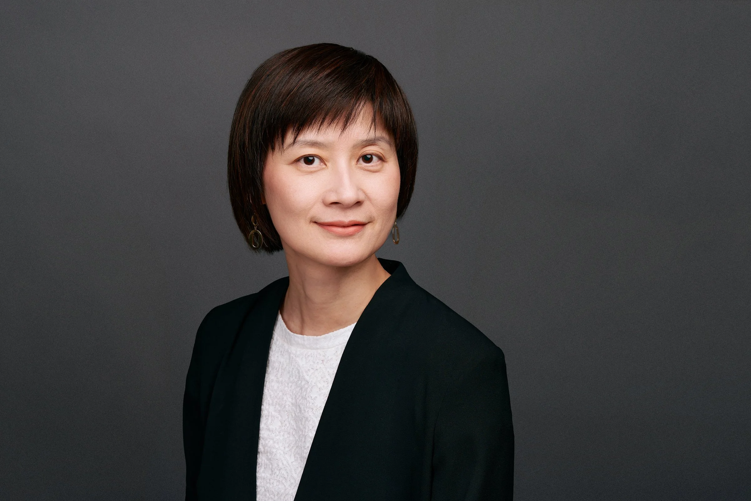 A corporate headshot of a professional woman with short dark hair wearing a black blazer over a white top, standing against a gray background.
