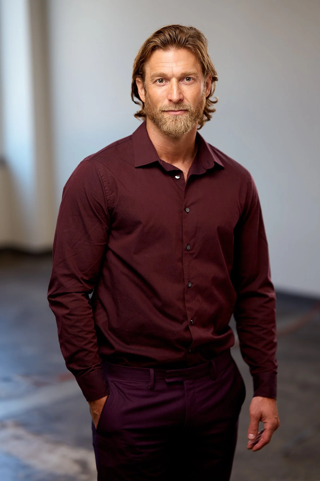 A man with shoulder-length brown hair and a beard, wearing a burgundy dress shirt and dark pants, standing indoors with one hand in his pocket.