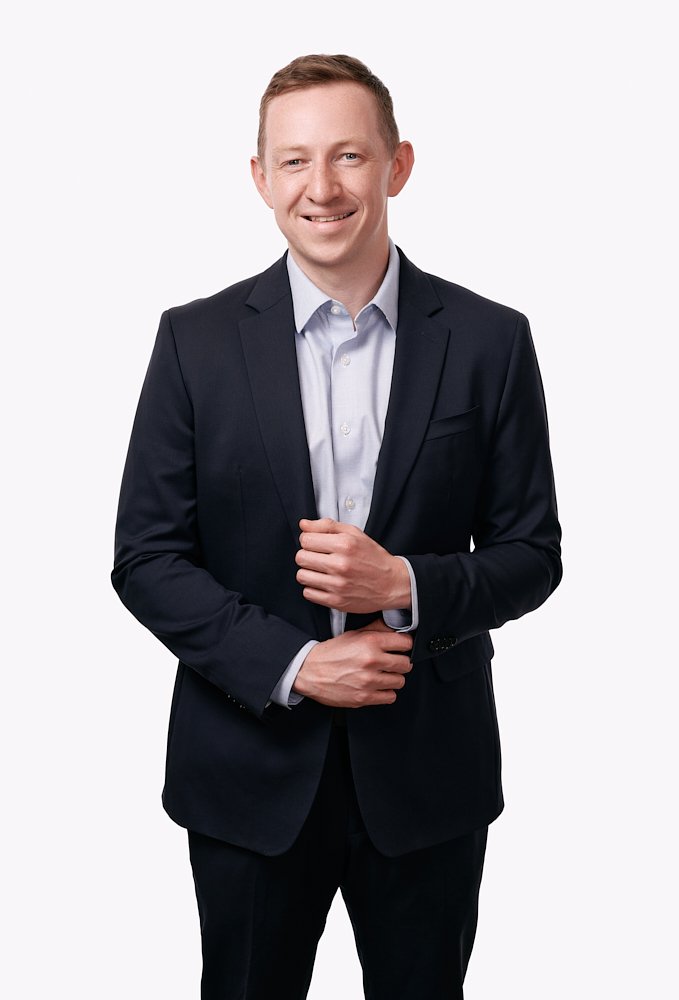 Professionally dressed man in a dark suit and light blue shirt, smiling, standing against a white background.