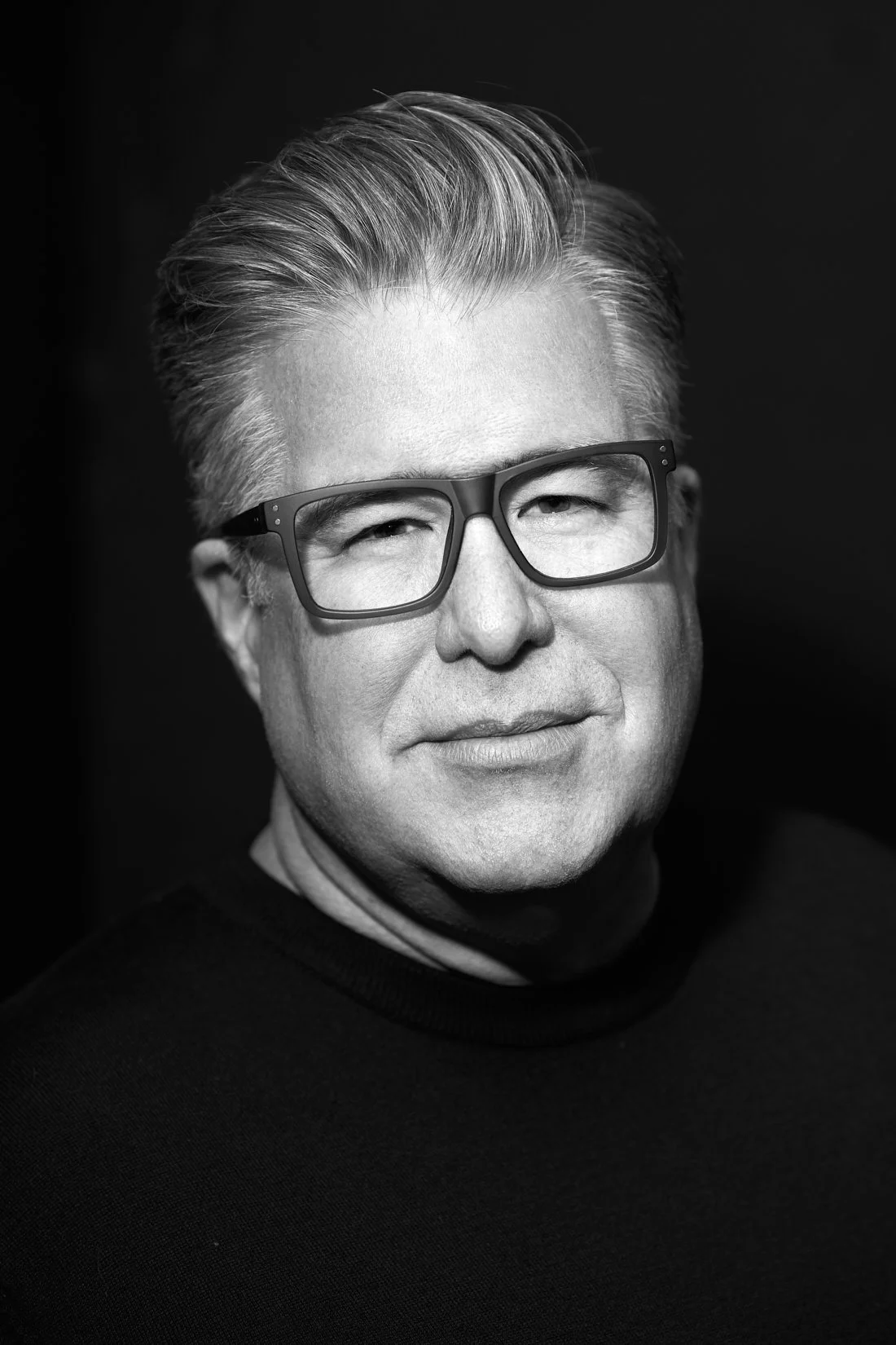 Black and white portrait of a middle-aged man with short, styled hair, wearing glasses and a dark turtleneck sweater, against a solid dark background.