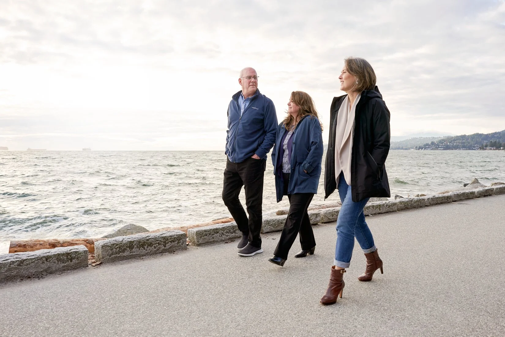 Three people walking along a waterfront promenade, with the ocean behind them and a distant landmass to the right, wearing jackets and casual attire.