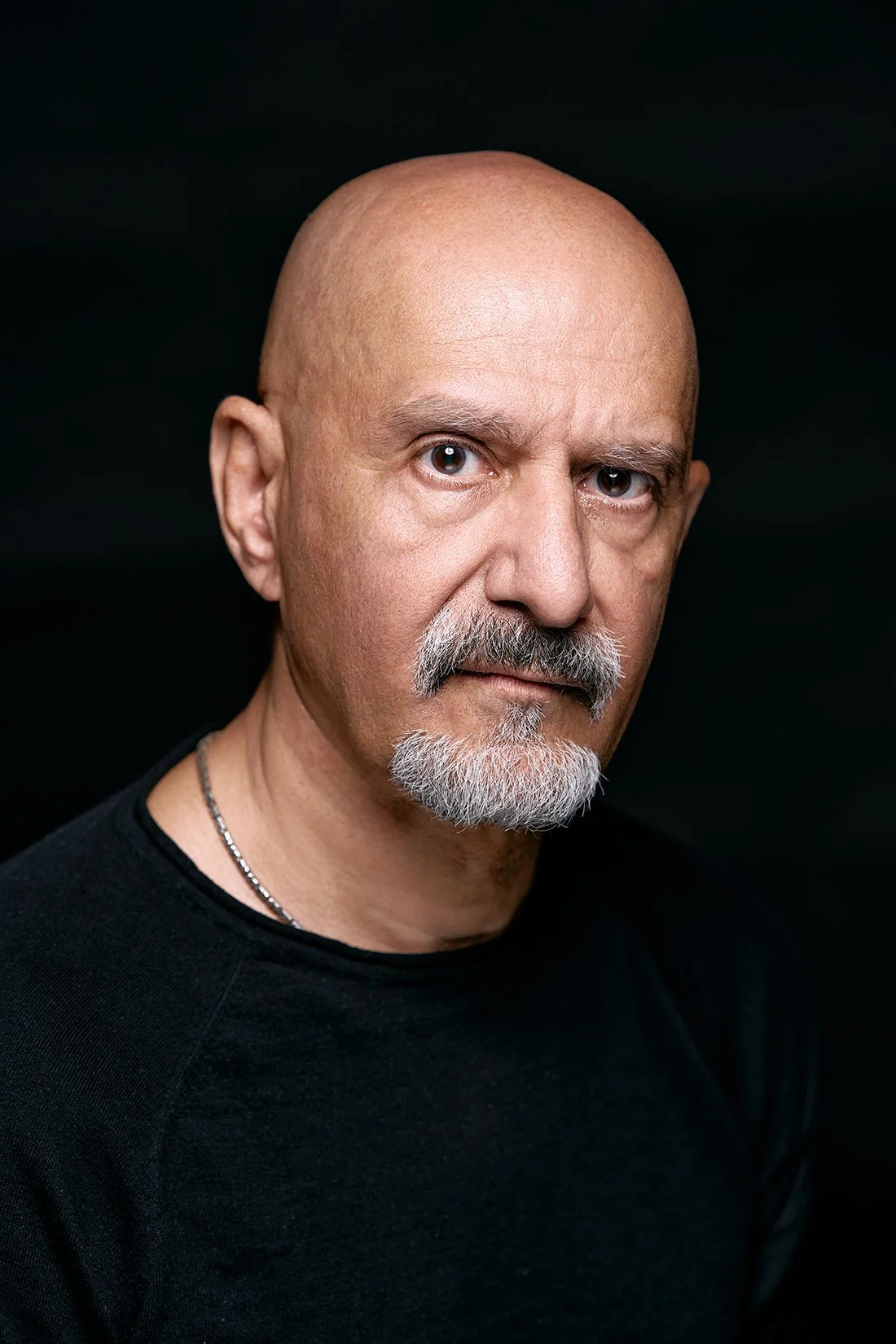 Close-up of a bald man with a gray beard and mustache, wearing a black shirt and necklace, against a dark background.