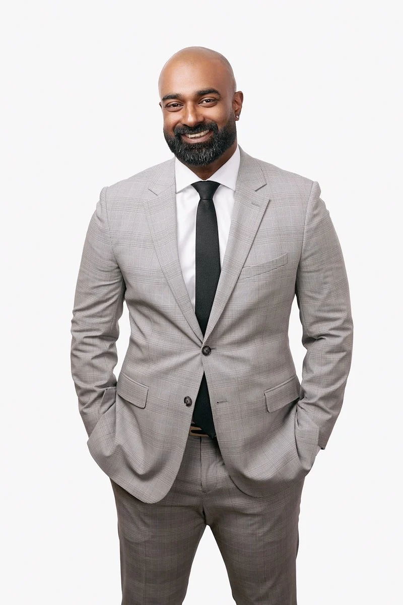 A branding portrait of a smiling man with a bald head, beard, and earrings wearing a light gray suit with a black tie, standing against a plain white background.