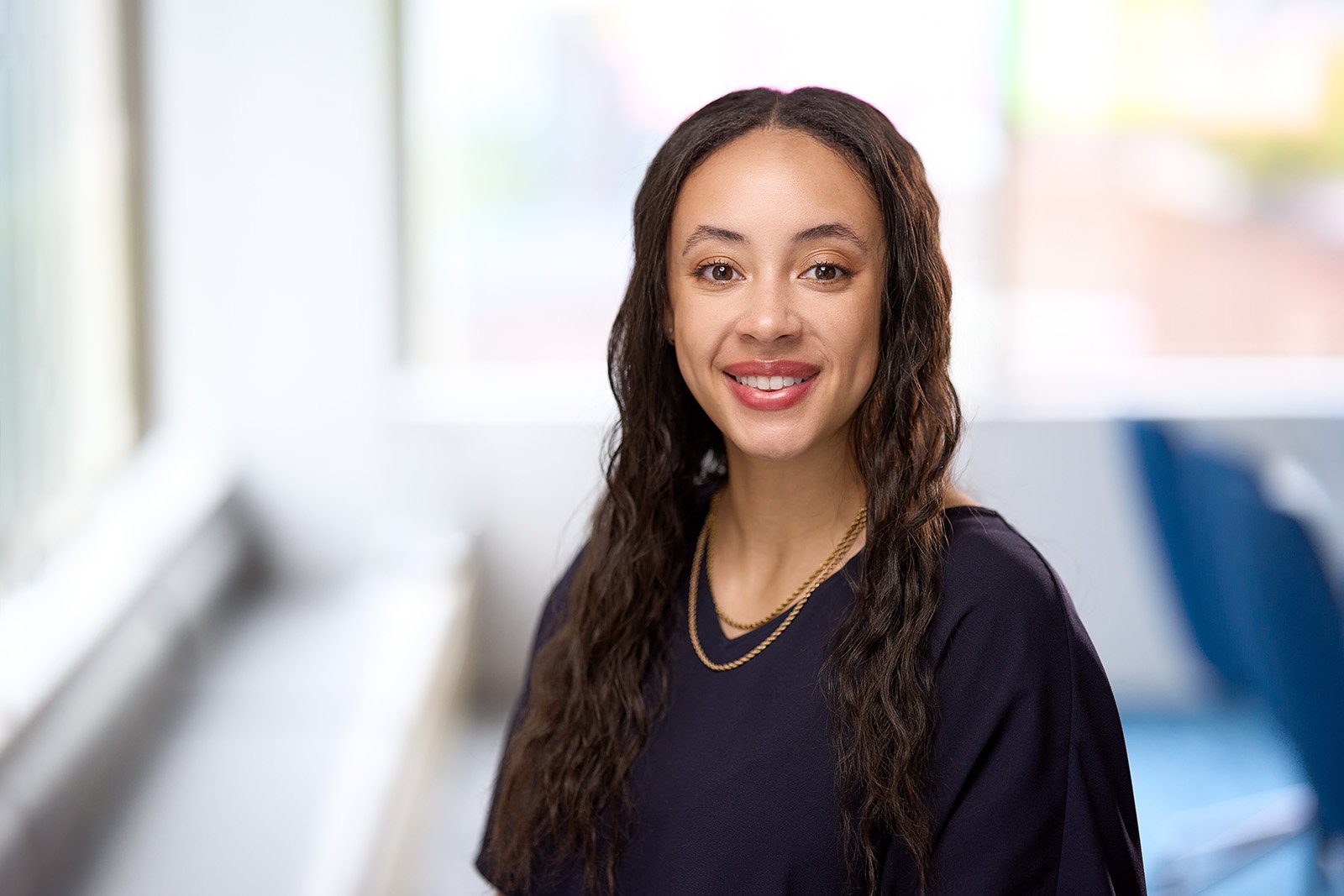 Corporate headshot of a woman with long wavy dark hair smiling, wearing a black top and layered gold necklaces, sitting indoors near a window with a blurred background.