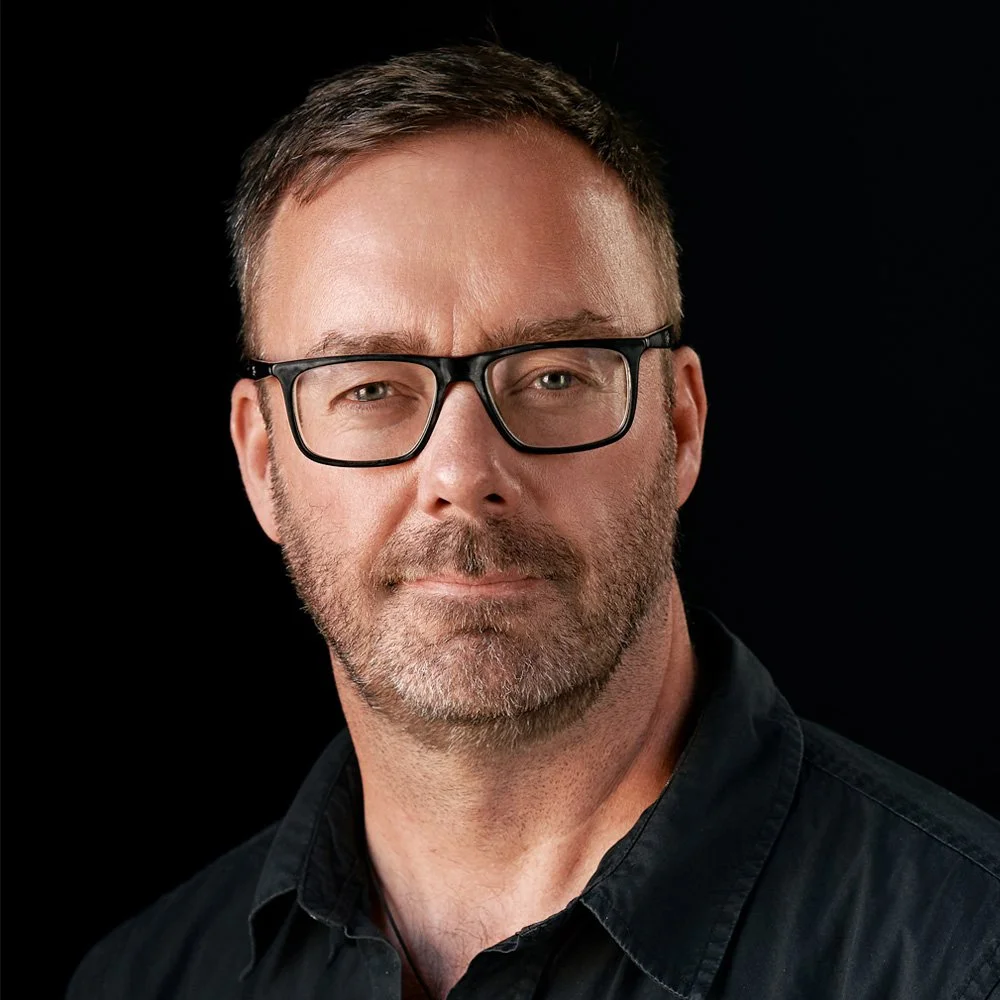 A man wearing glasses with short brown hair, a beard, and a black shirt, posing against a dark background.