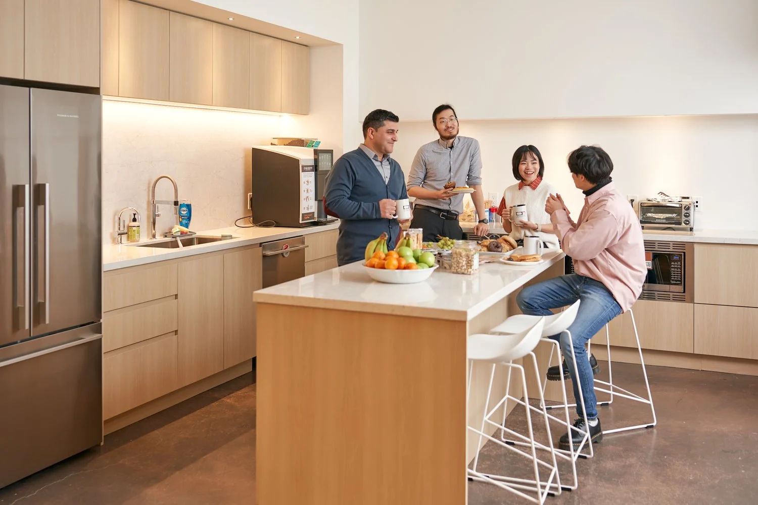 Group of five people socializing in a modern kitchen, holding mugs, with breakfast foods and fruits on the countertop.