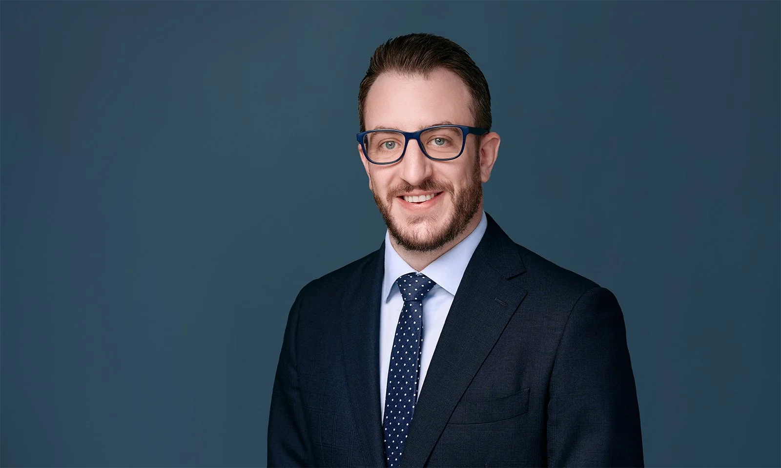 Corporate headshot of a man in a dark suit, light blue shirt, and polka dot tie smiling against a plain dark background.
