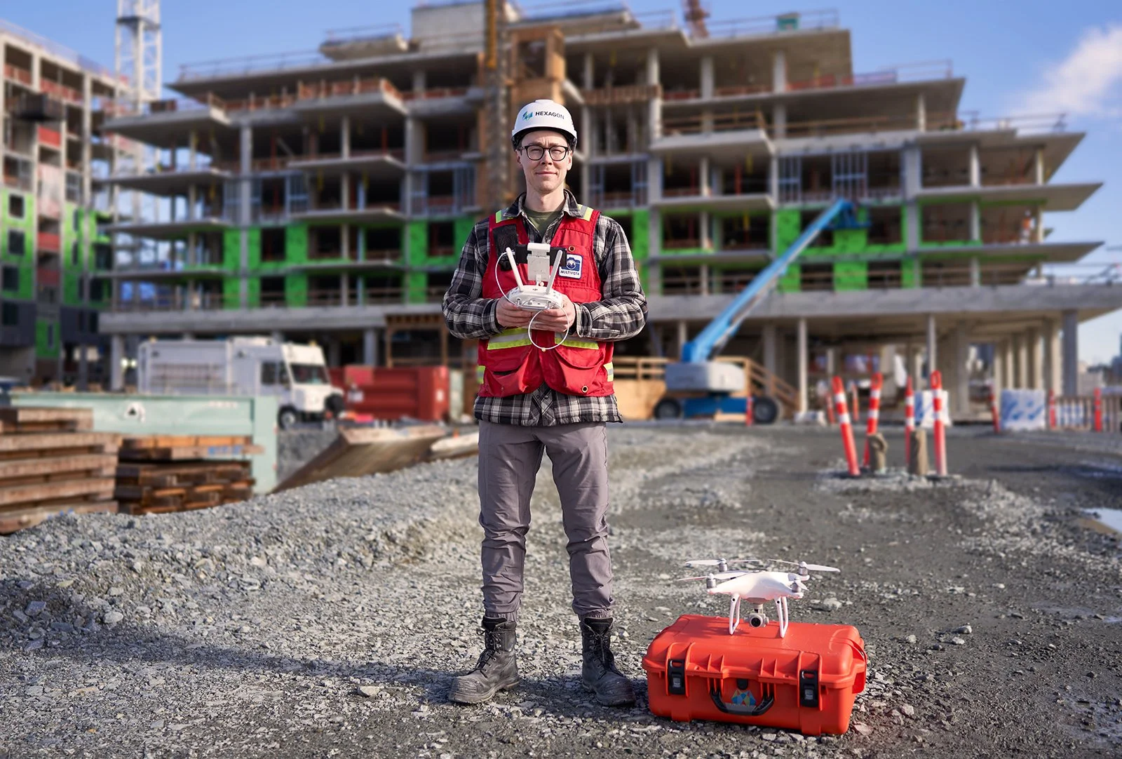 Construction worker standing on a gravel site controlling a drone with a remote, with a large multi-story building under construction in the background.