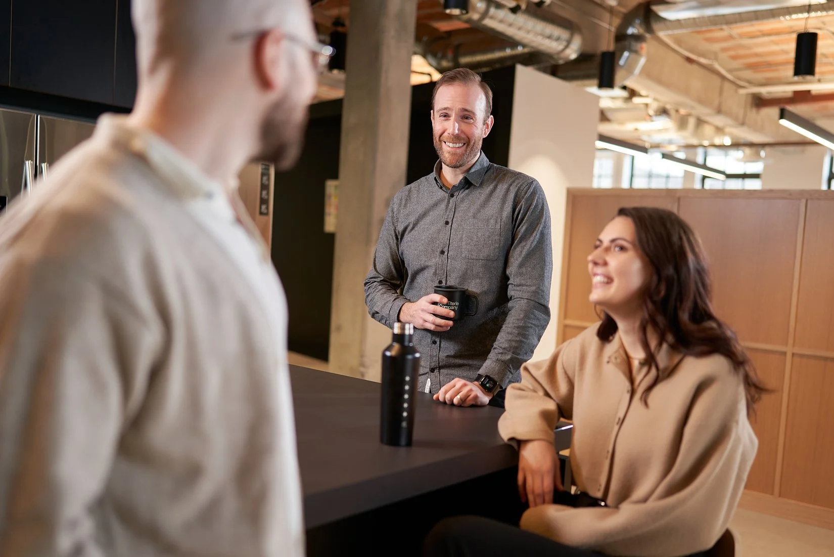 Three people having a conversation in an office kitchen; a woman sitting at a counter, a man standing with a coffee mug, and another man with blurred features in the foreground.