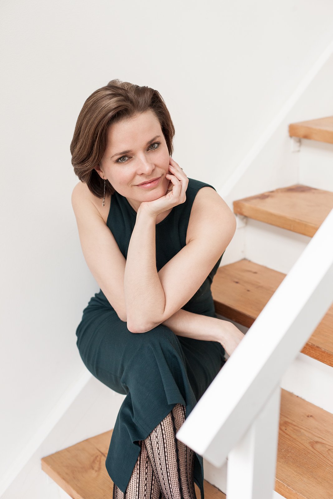 A woman with short brown hair and a sleeveless dark green dress sitting on wooden stairs, resting her chin on her hand, looking at the camera.