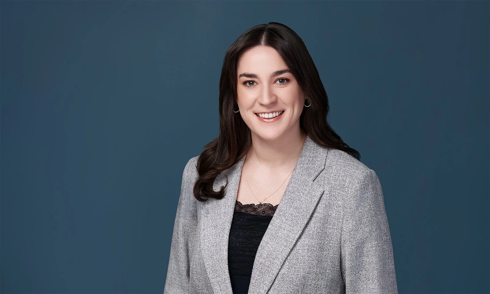 Headshot of a woman with long dark hair smiling, wearing a light gray blazer over a black lace top, against a blue background.