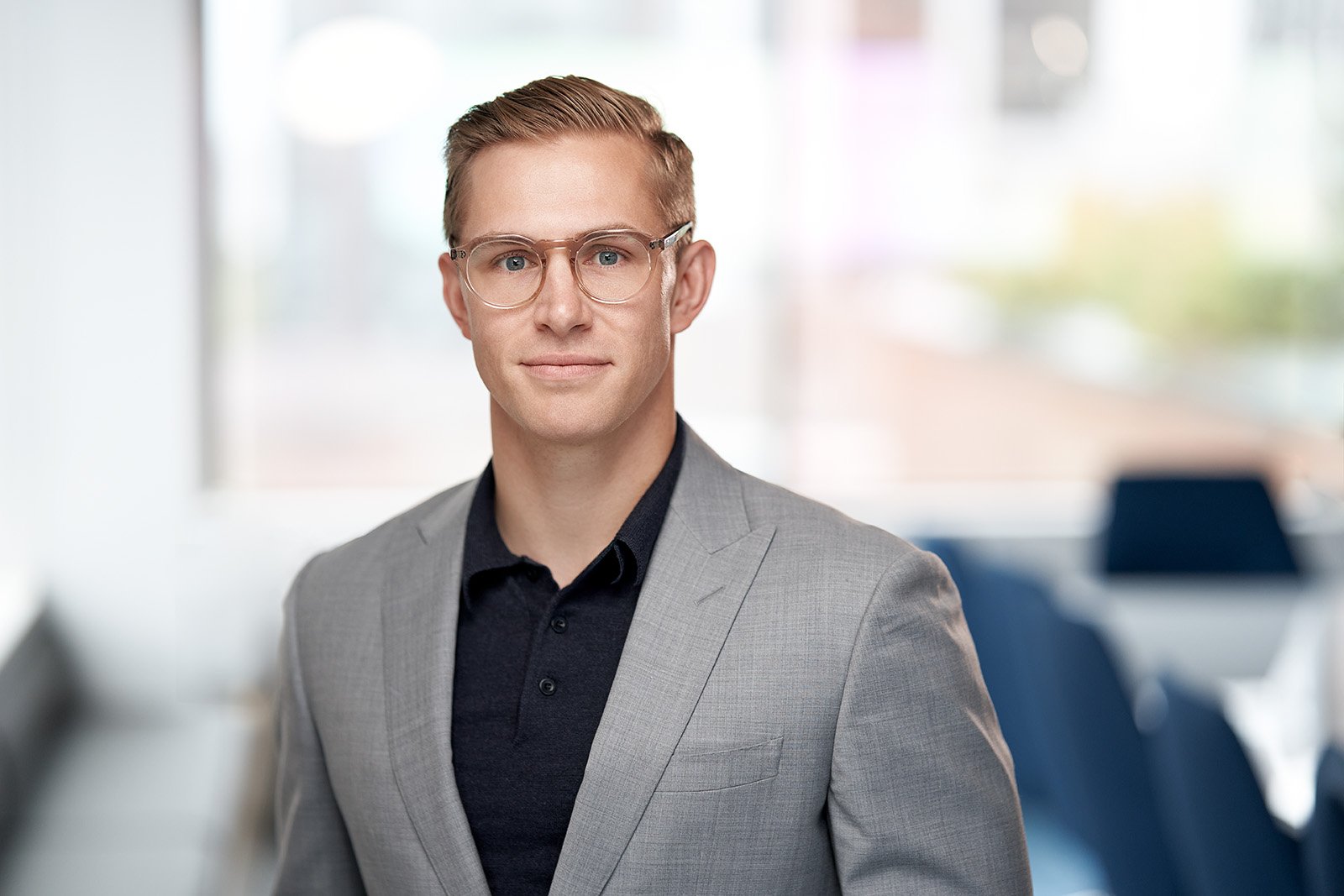 Corporate headshot of a young man with blonde hair, glasses, wearing a light gray suit jacket and black shirt, standing in a modern office with blurred blue chairs in the background.