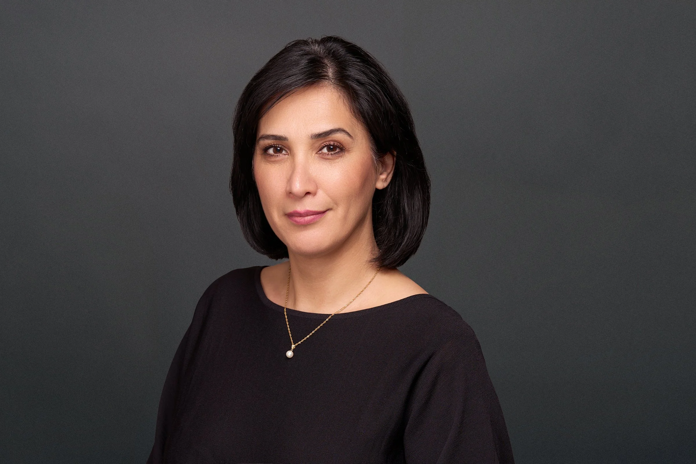 Corporate headshot of a professional woman with short black hair, wearing a black top and a gold necklace with a pearl pendant, posing against a dark gray background.