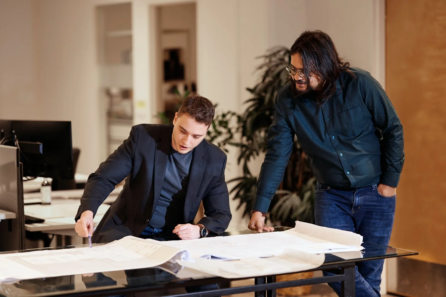 Two men in a modern office reviewing large architectural blueprints on a glass table, with computers and office supplies in the background.