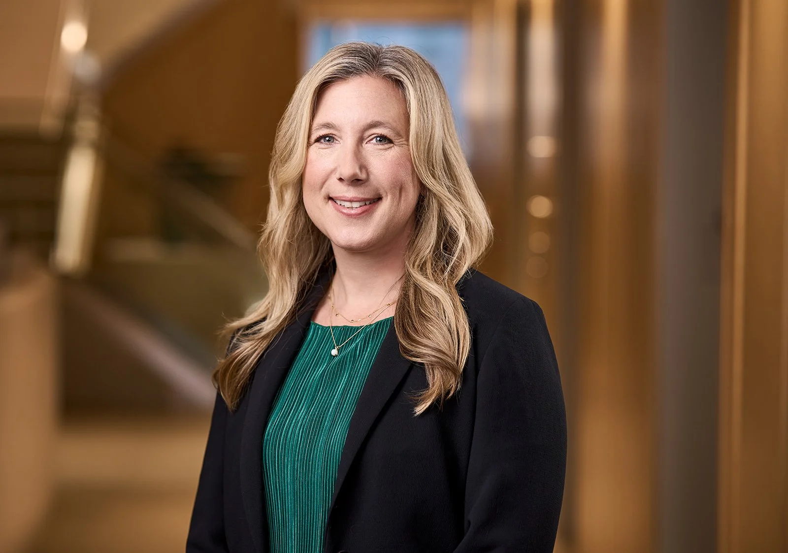 A corporate portrait of a professional woman with long blonde hair, wearing a black blazer and a green top, smiling in an indoor setting with warm lighting and wooden decor.