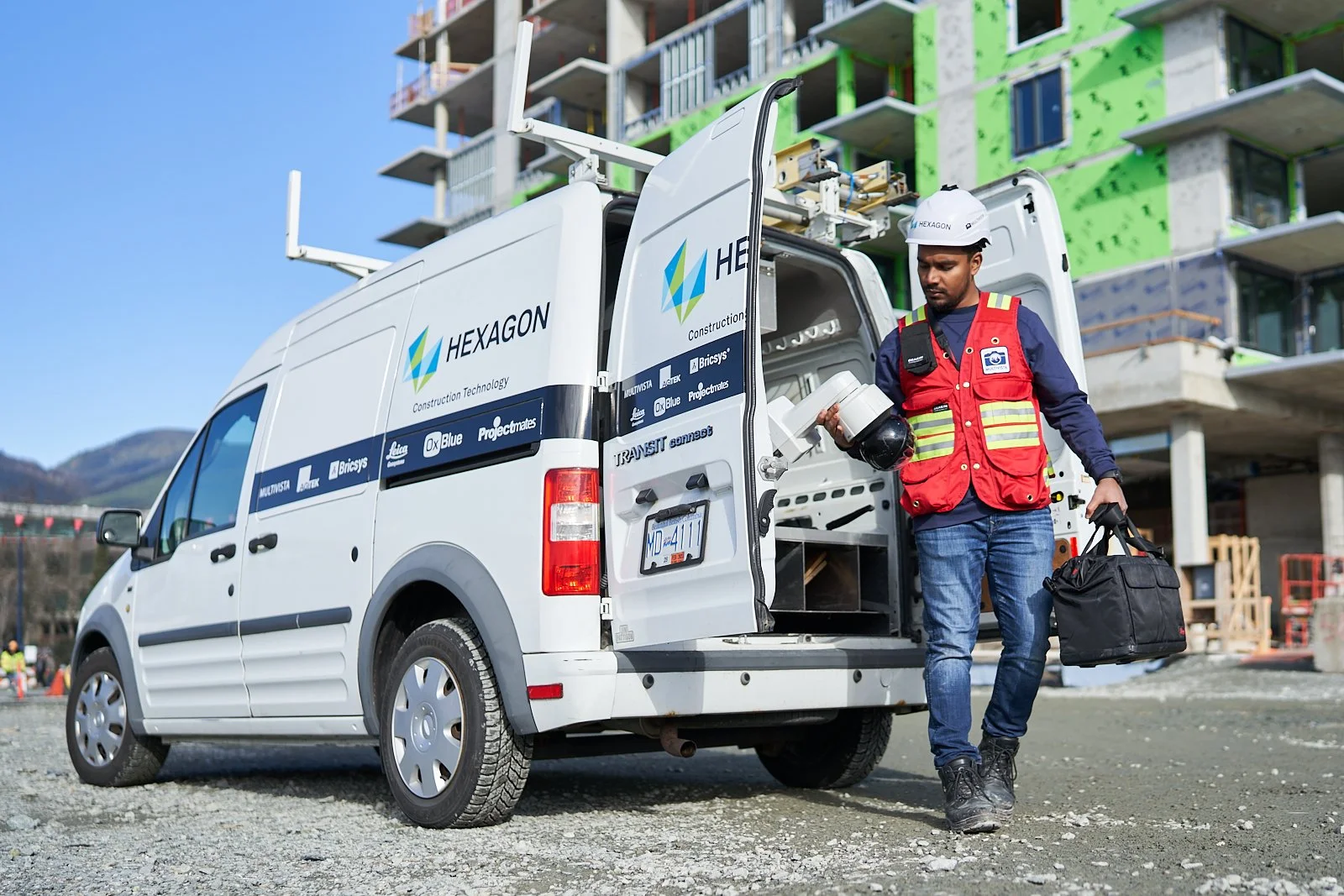 Construction worker unloading equipment from a white Hexagon construction van at a building site with an under-construction building in the background.