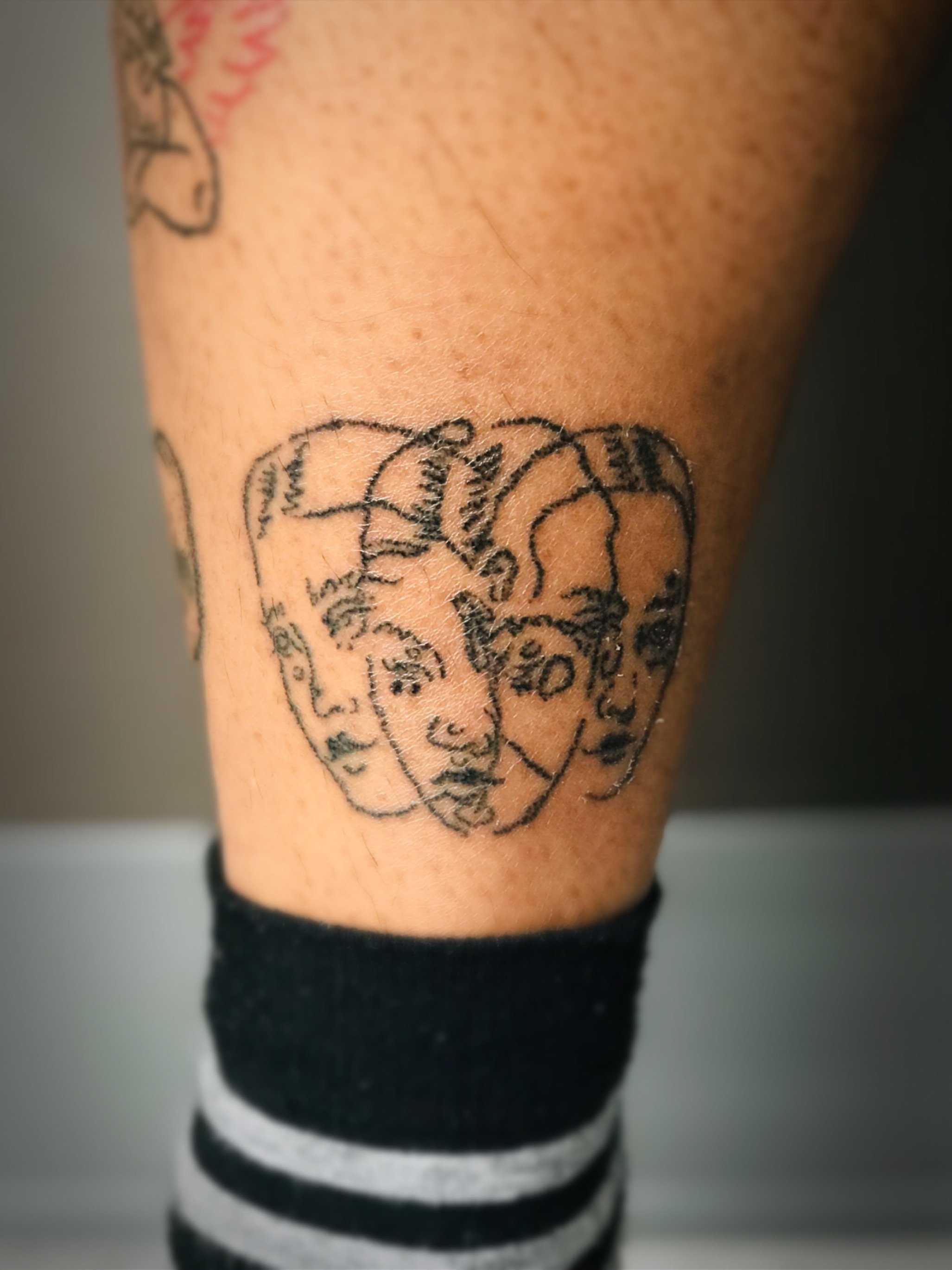Tattoo of three overlapping abstract faces on a person's leg, with a black and gray striped sock below.