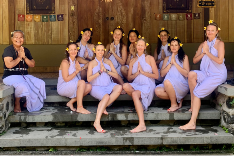 A group of ten women in lavender dresses and yellow flower crowns sitting and standing on steps in a wooden temple, smiling with hands in prayer position.