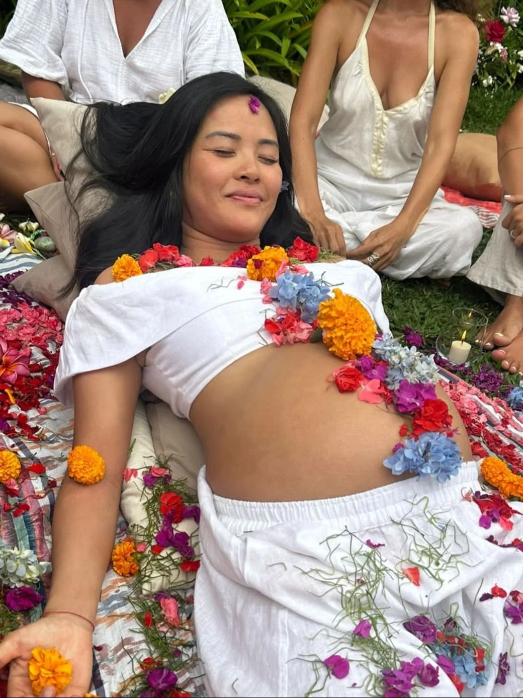 A pregnant woman lying on her back surrounded by colorful flowers, with a serene expression. Several women sit around her in white clothing, in an outdoor garden setting.
