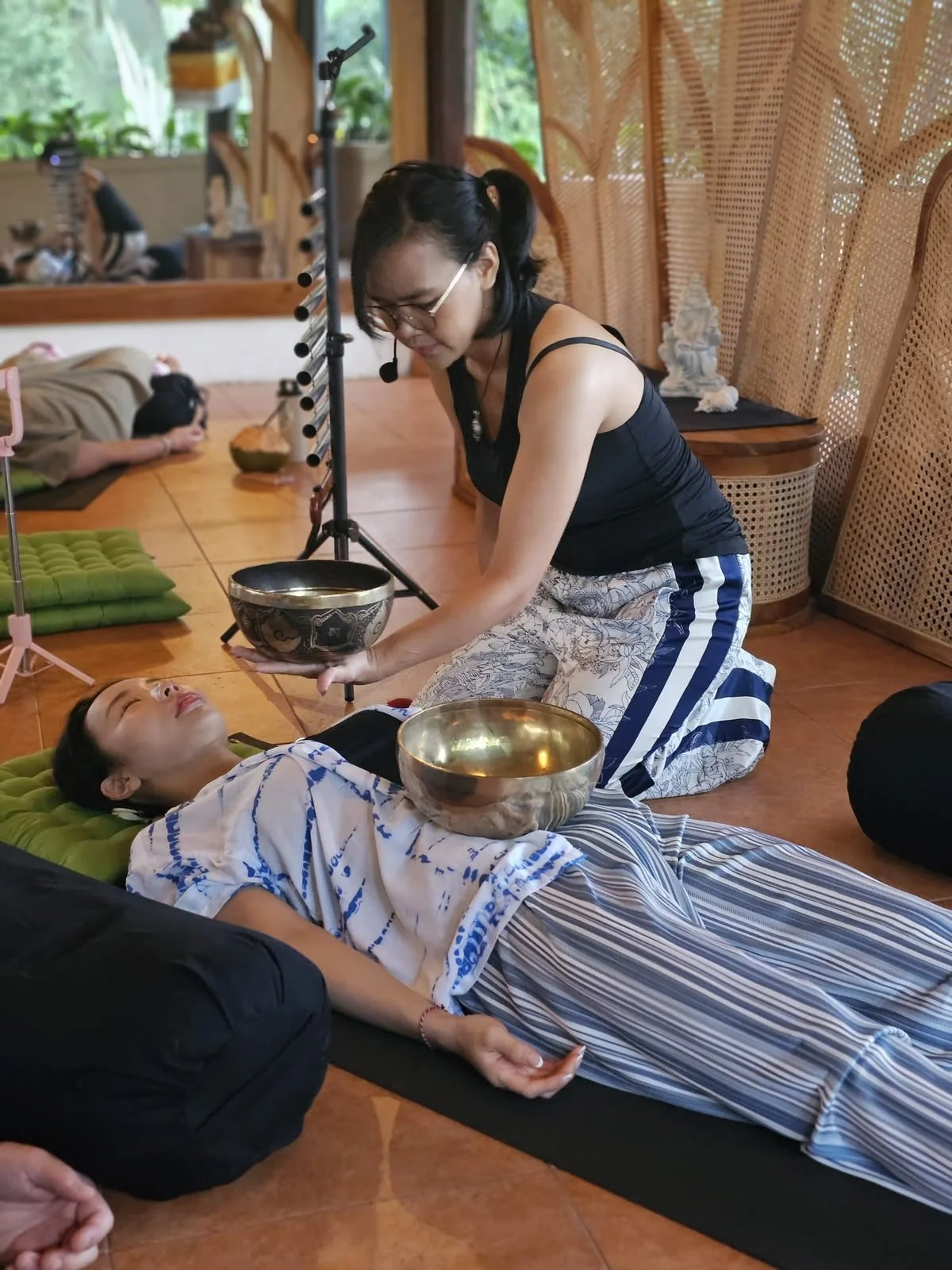 A woman performing a sound healing or meditation session on a person lying on the floor, with singing bowls and a mallet in a room decorated with wooden furniture and natural light.