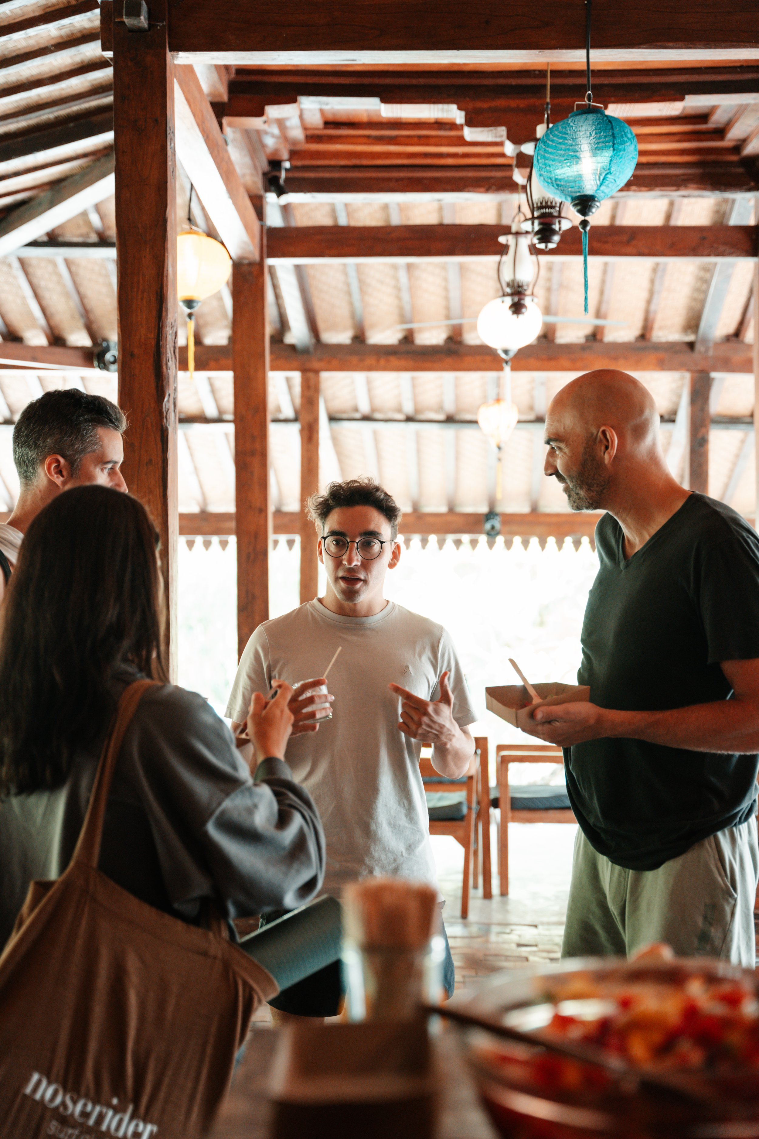Group of people sitting on the floor inside a rustic wooden building, engaged in a discussion or workshop. There are papers and materials scattered around. The environment appears natural and tropical with greenery visible outside.