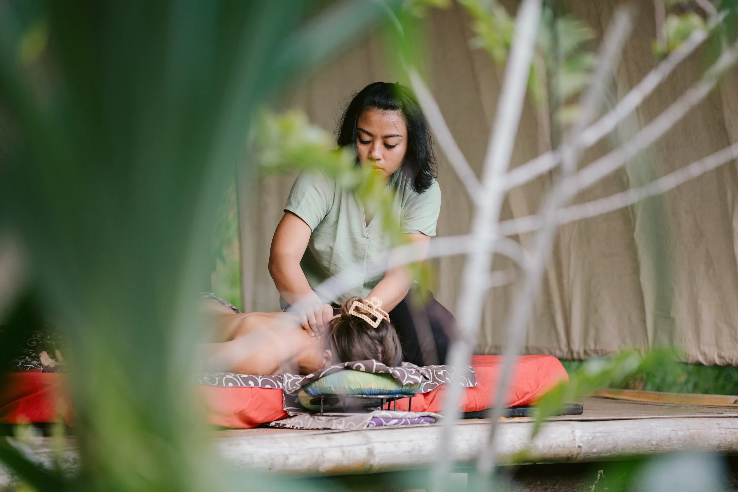 A woman receiving a massage outdoors on a red cushioned mat, surrounded by greenery and plants.