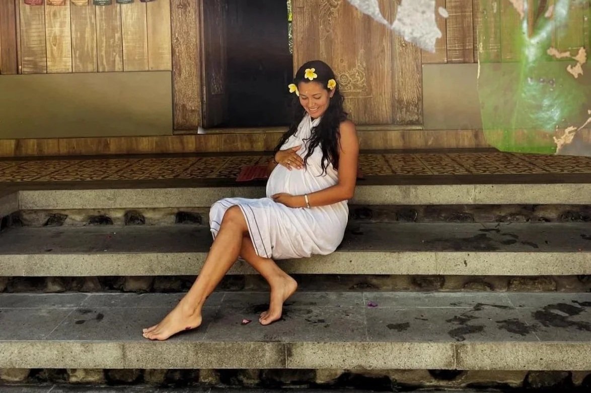 A pregnant woman sitting on concrete steps, smiling and touching her belly, wearing a white dress and a flower crown.