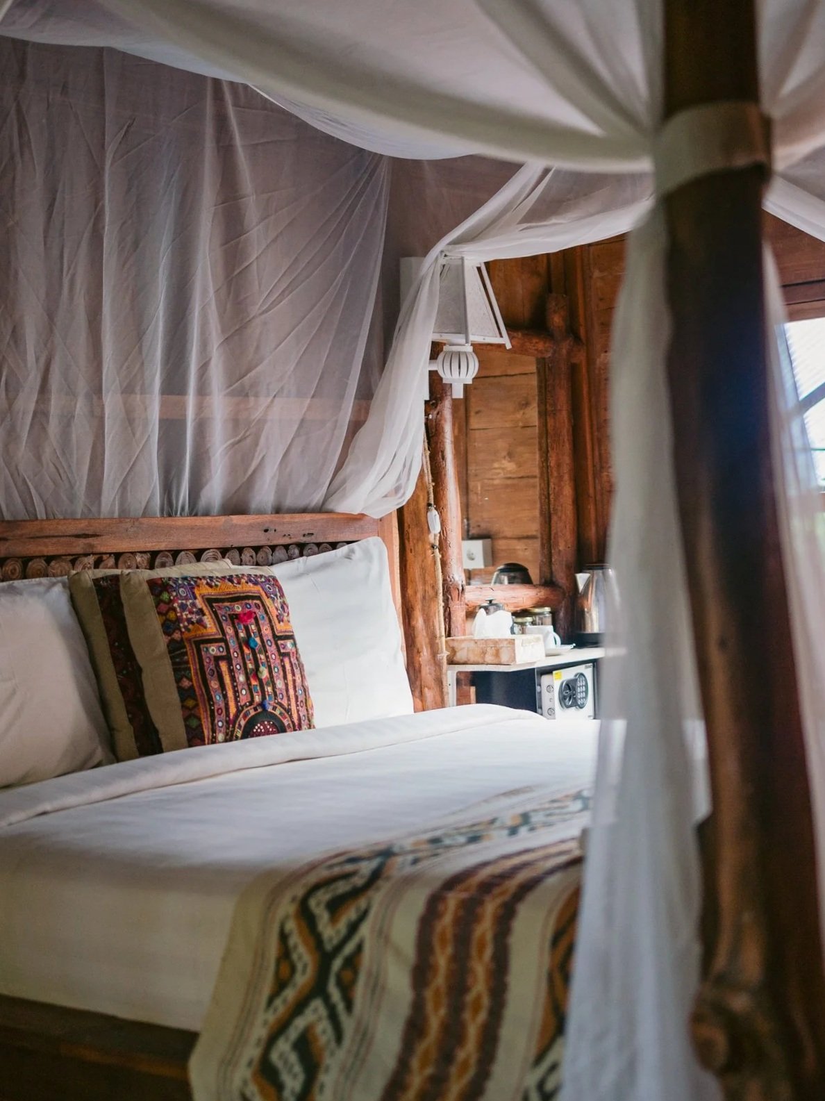 Inside a rustic bedroom with wooden walls and furniture. A bed with white sheets, a colorful embroidered pillow, and a patterned blanket. There is a mosquito net over the bed and a side table with various appliances.