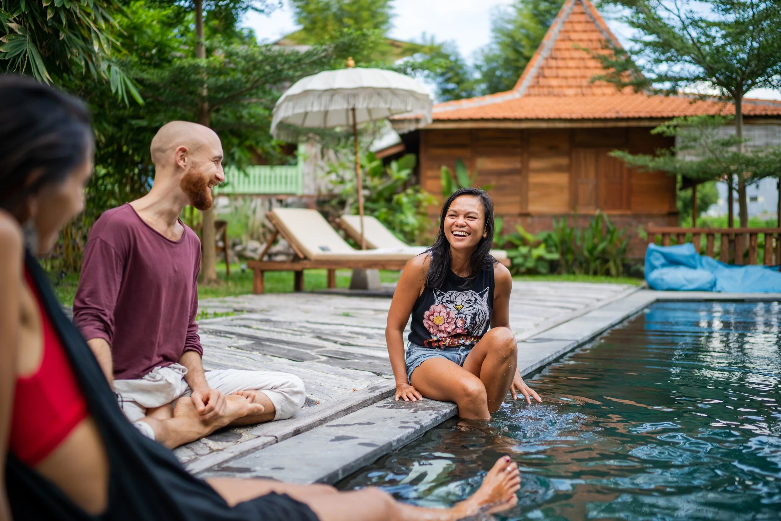 Group of four friends sitting by a pool in a backyard, smiling and laughing. One woman is sitting on the edge with her feet in the water, while the others are in the background. There is a wooden house with a red tile roof, green trees, and a white umbrella in the yard.