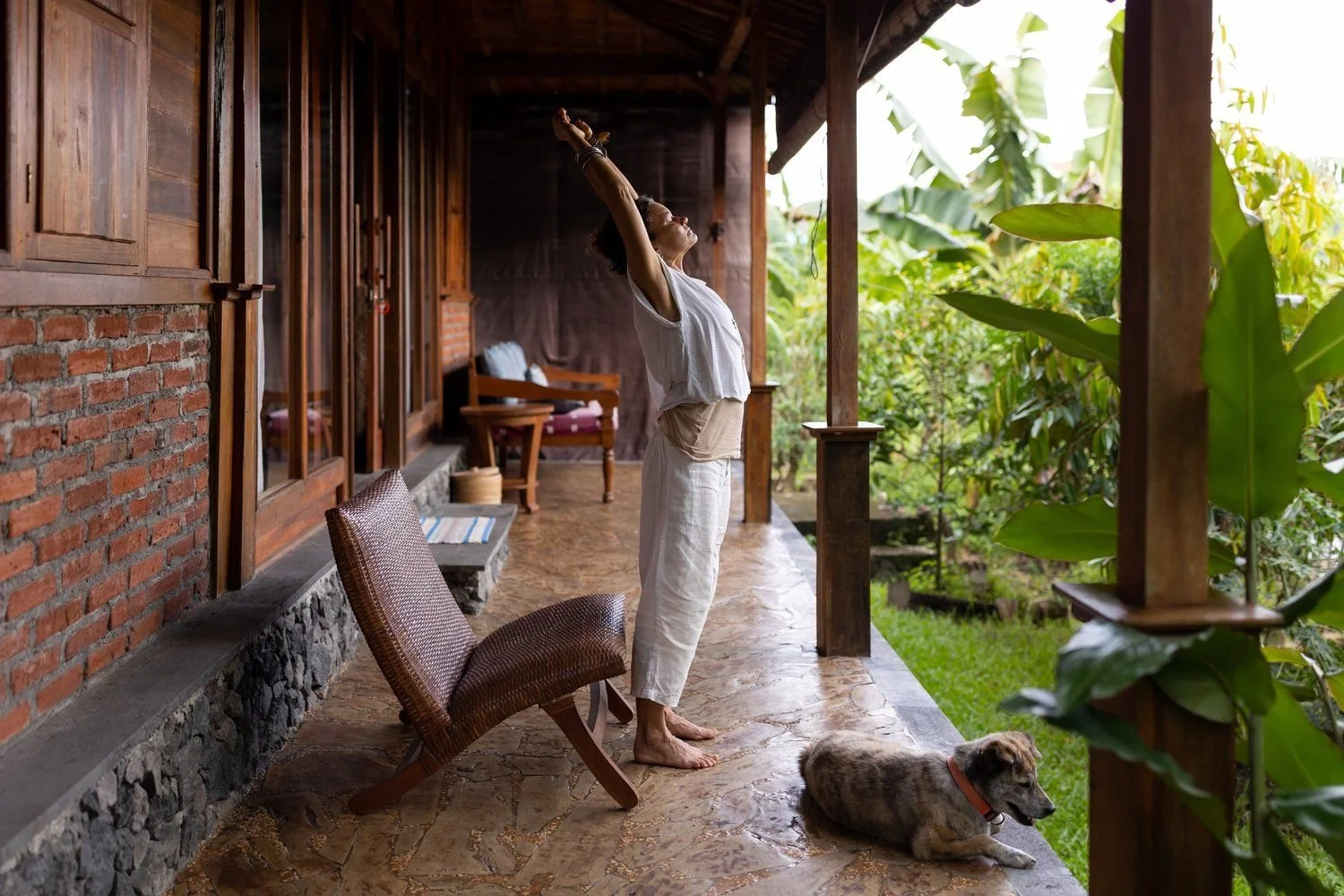 A woman in white clothing stretching on a porch next to a resting dog, with lush green plants outside.