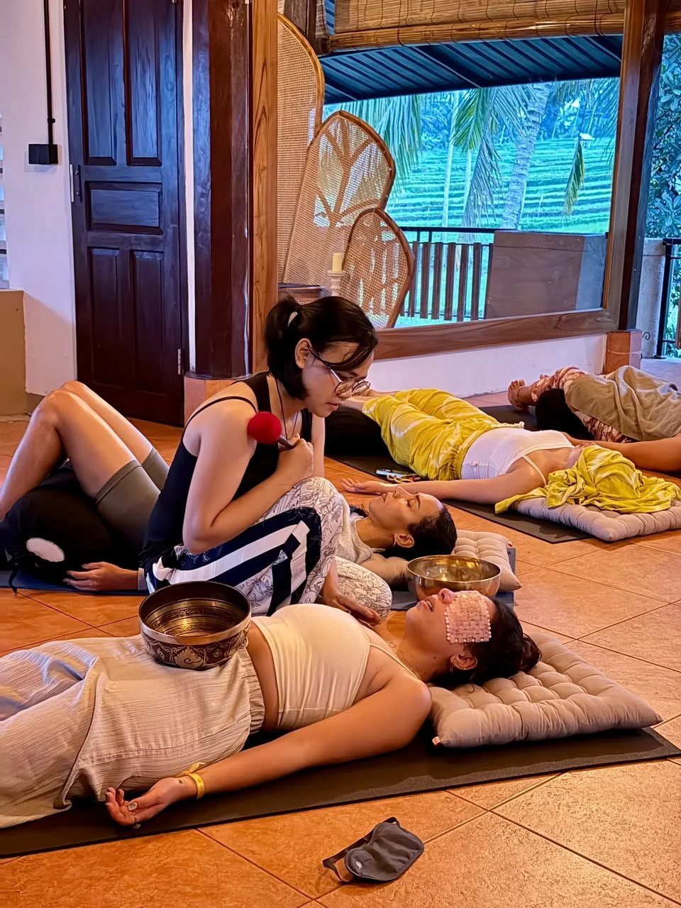 Group of people lying on yoga mats, participating in a relaxation or meditation session inside a room with wooden elements and large windows showing tropical trees outside.