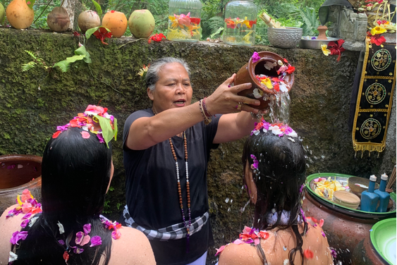 An elderly woman pouring water over two young women during a traditional water blessing ceremony, with flower petals on their heads and various decorative items around them.
