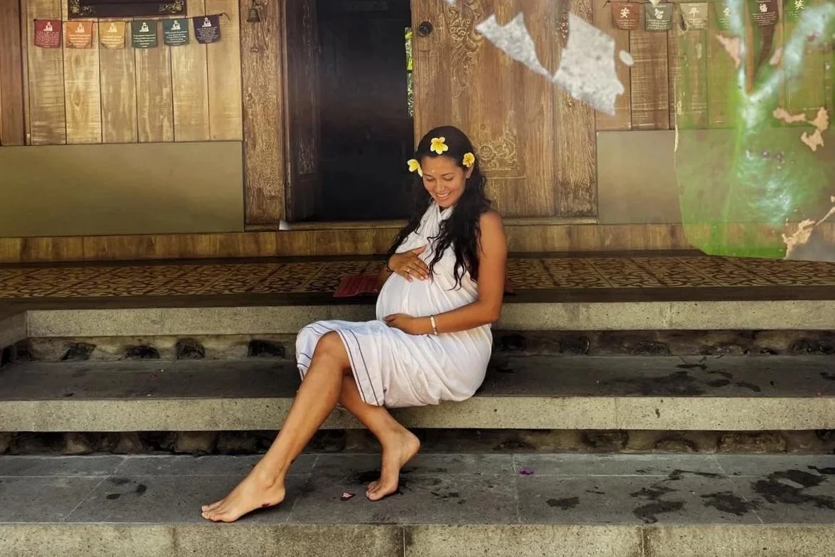 A pregnant woman with long dark hair, wearing a white dress and a flower crown, sitting barefoot on stone steps in a wooden setting, smiling and holding her belly with one hand.