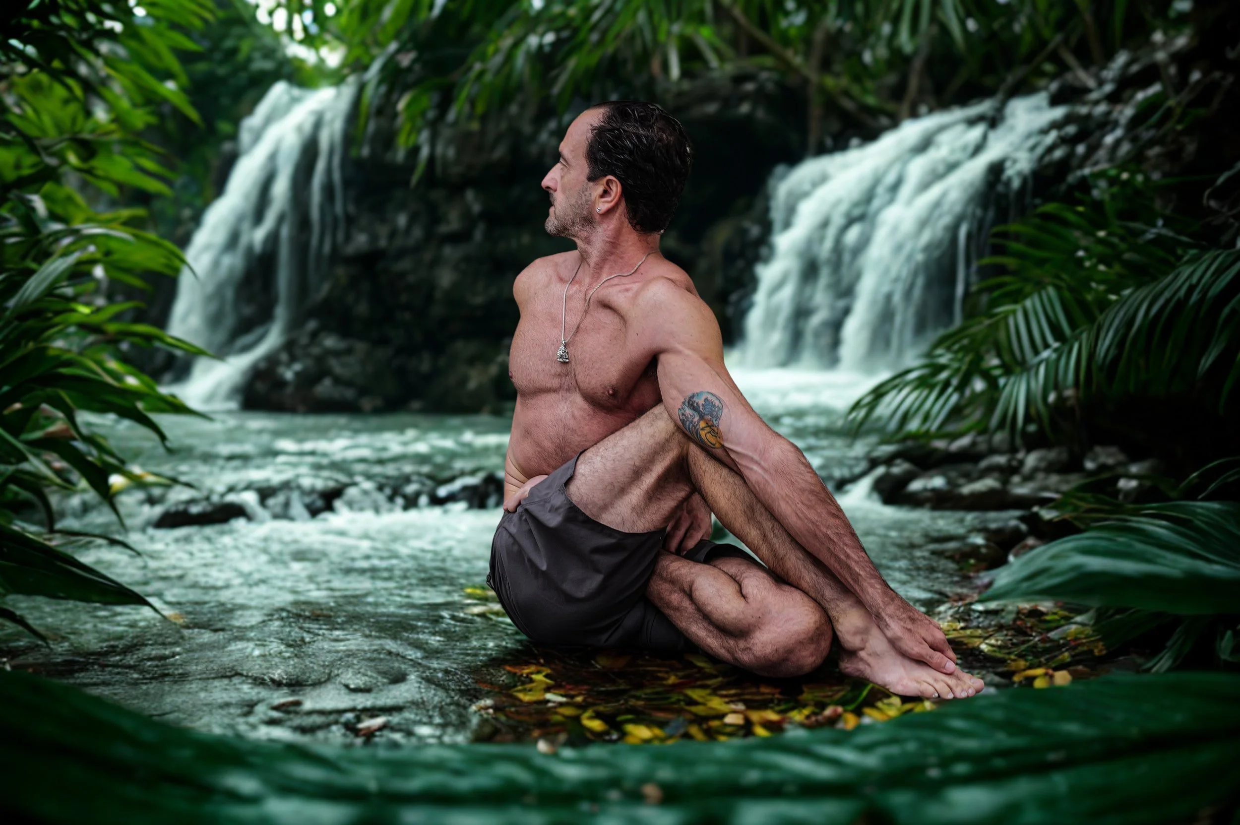 A shirtless man with tattoos on his arm and gray shorts is practicing yoga in a kneeling position on a rock in a river, with lush green foliage and a waterfall in the background.