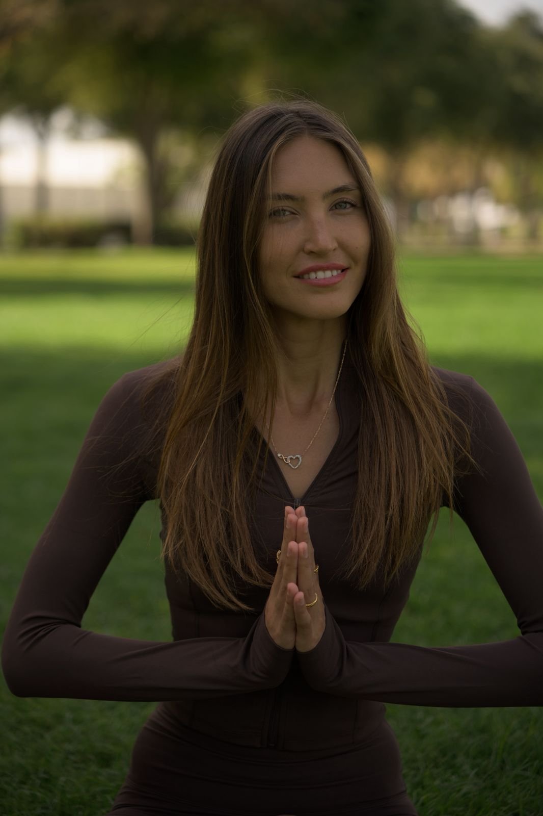 A woman doing yoga outdoors in a park with trees in the background, standing in a prayer pose with her hands together in front of her chest and smiling.