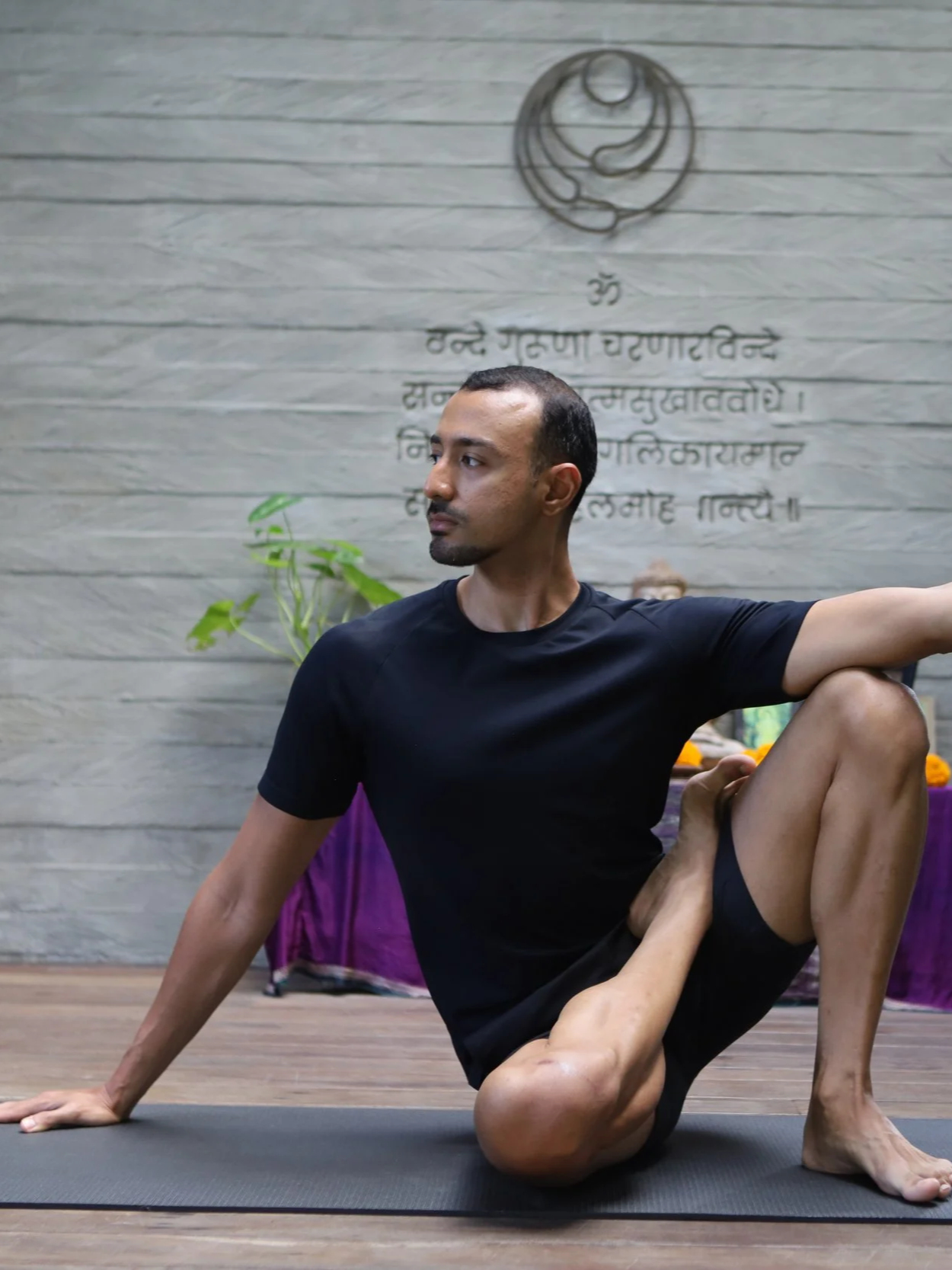 A man practicing yoga indoors, performing a pose on a yoga mat with a brick wall and a spiritual symbol with text behind him.