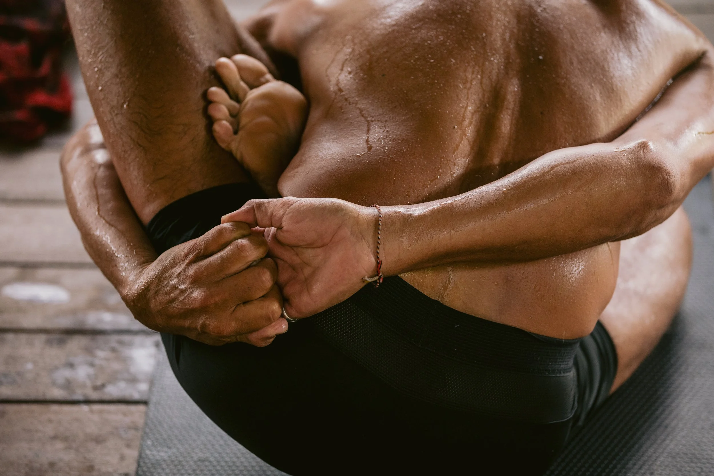 Close-up of a shirtless man with a muscular build sitting on a gray exercise mat, holding his knee during a workout.