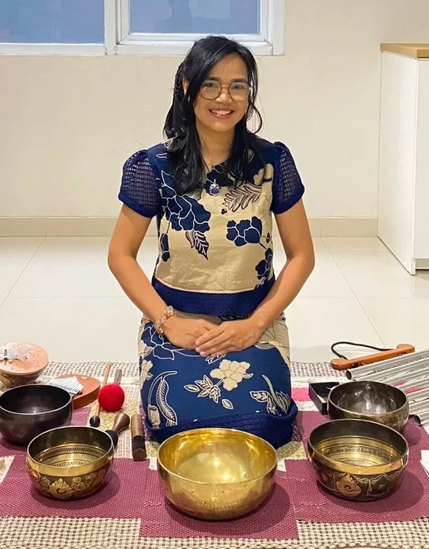 A woman wearing glasses and a blue and beige floral dress sits on the floor in front of a collection of singing bowls and mallets, smiling at the camera.