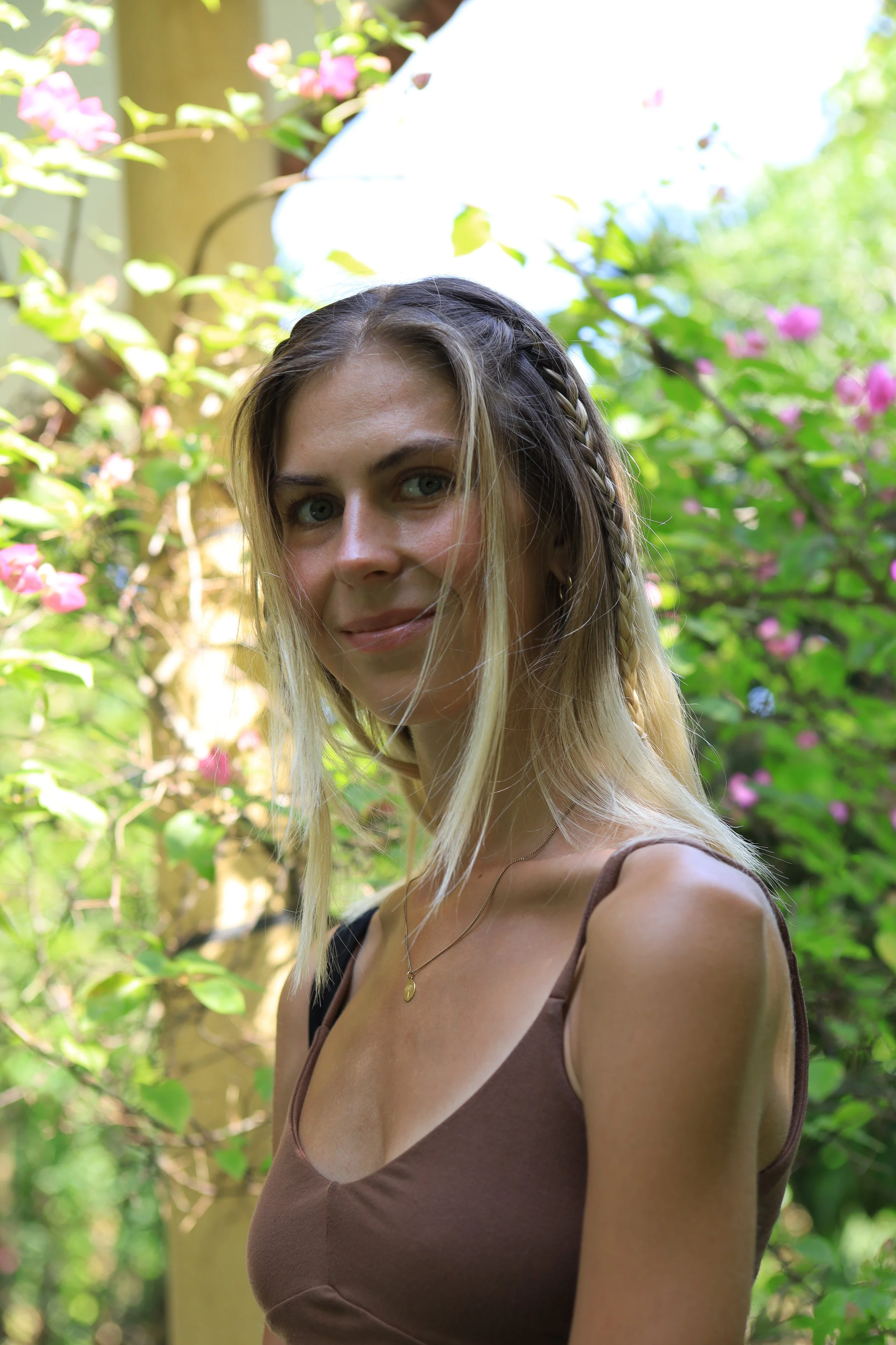 A young woman with blonde hair and blue eyes smiling outdoors, wearing a brown tank top and a gold necklace, with green trees and pink flowers in the background.