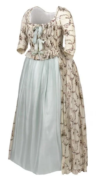A historical dress with a cream-colored bodice and sleeves, decorated with a brown floral pattern, featuring a ribbon at the neckline, paired with a white skirt that has a light blue hue, and a matching cream-colored floral apron or overlay.