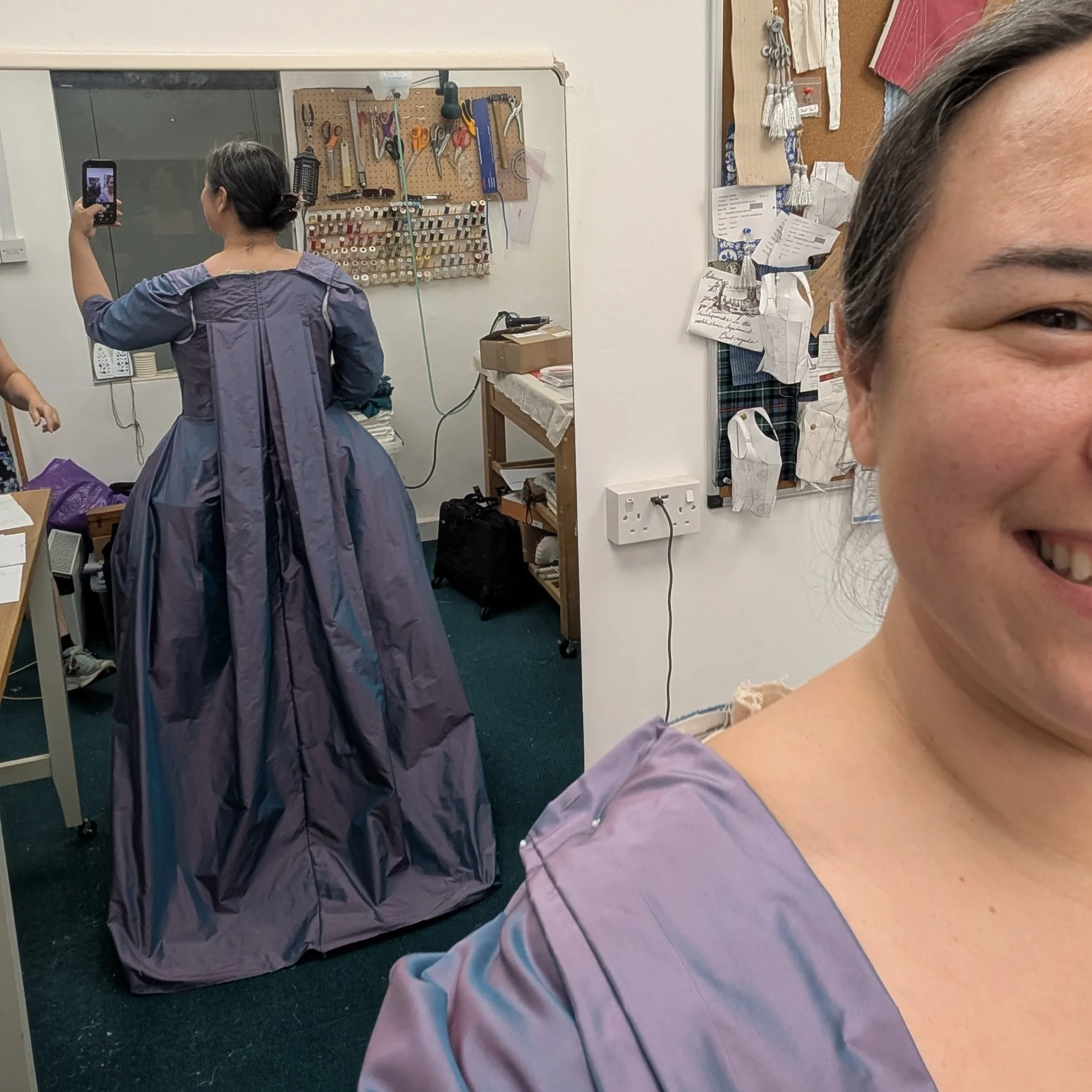 Person taking a mirror selfie in a workshop or craft room, showing a pegboard with various tools and a partial view of a smiling woman's face in the foreground.