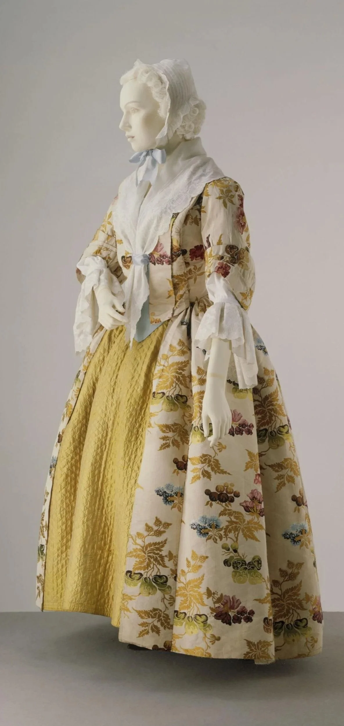 A mannequin dressed in a 18th-century style gown with floral and gold patterns, featuring a yellow skirt and a white lace blouse.
