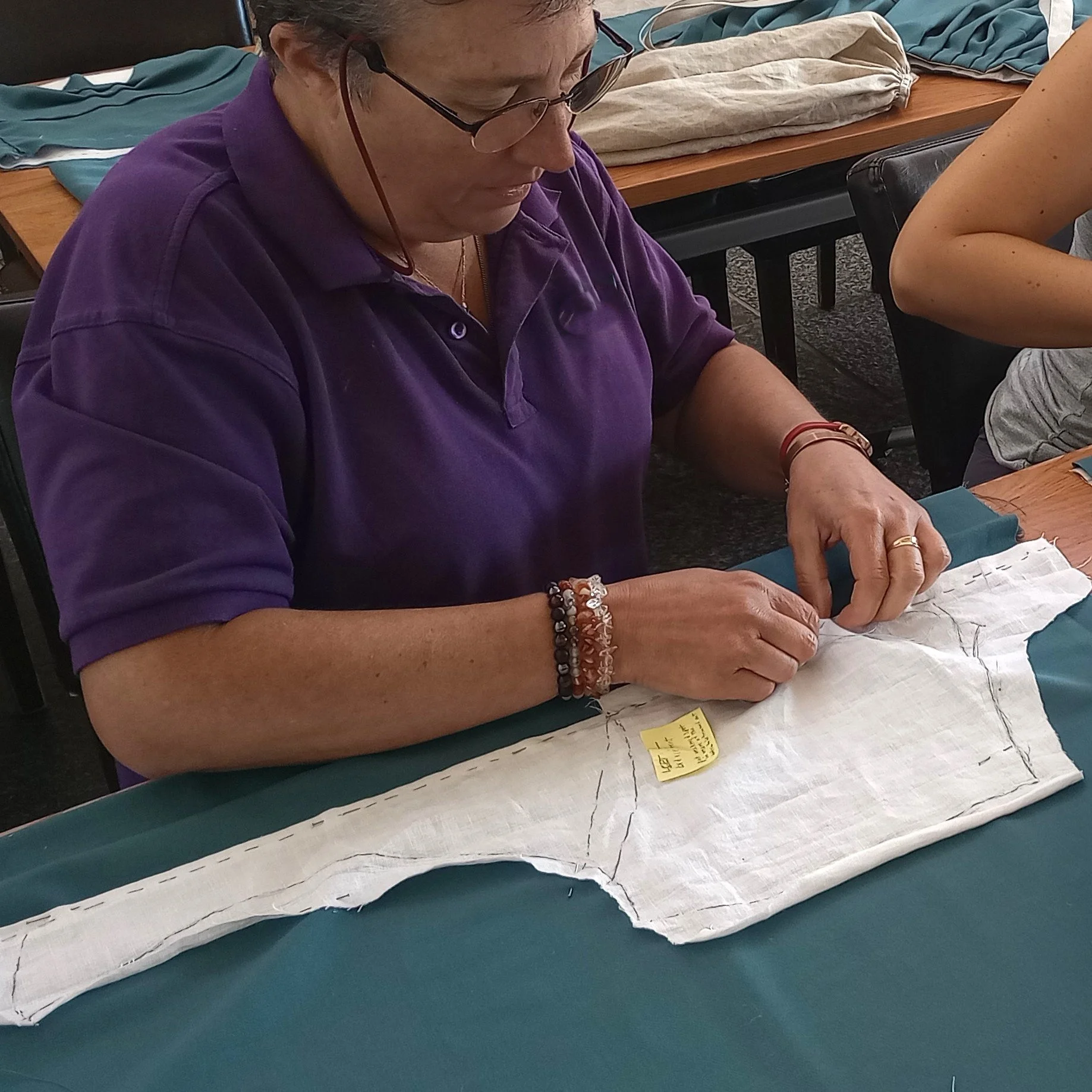 Tania builds the lining for one side of the bodice fronts, for the new 1740s English Gown she is making in one of Rebecca's workshops. Tania is wearing a purple polo shirt and spectacles.