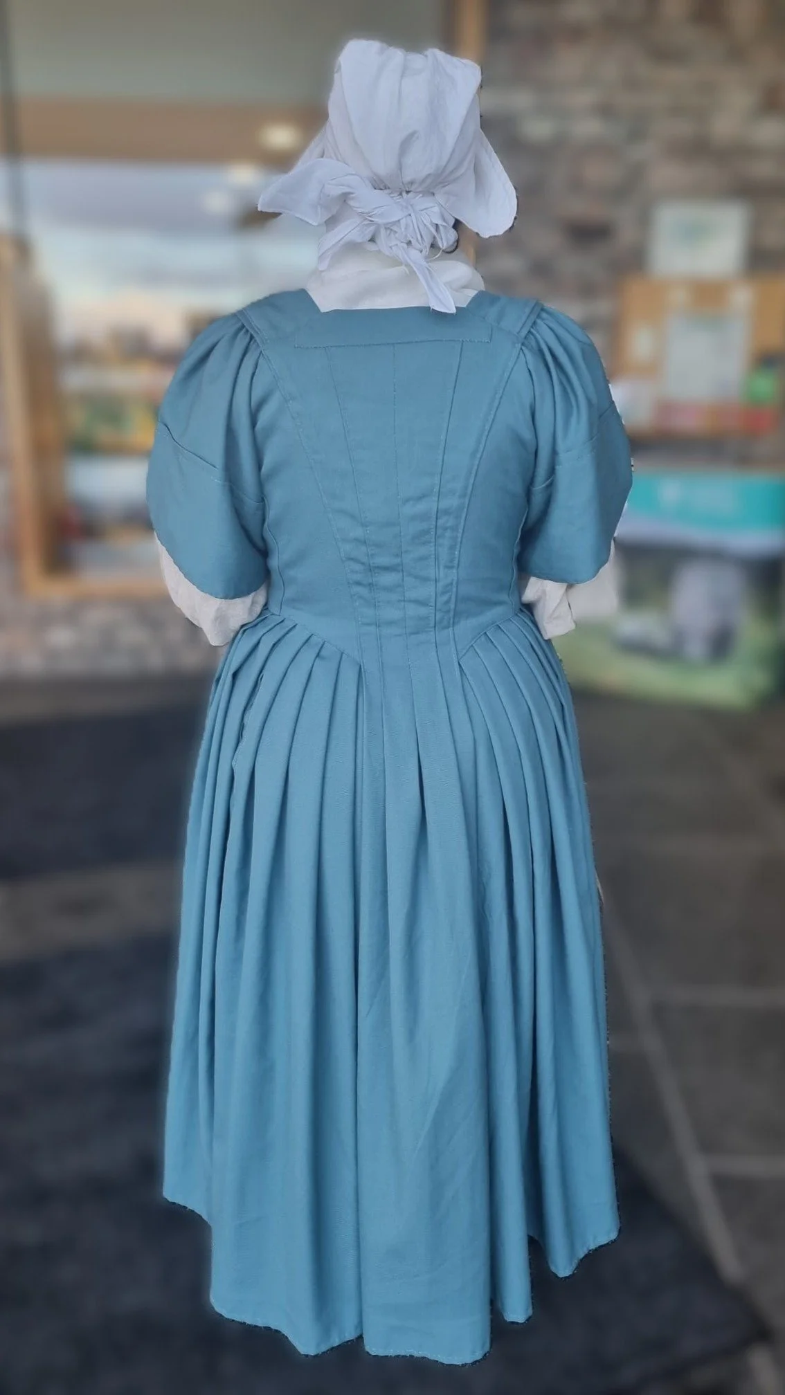 Tania modelling the new 1740s gown she made in one of Rebecca's gown-making workshops.  Her gown is made of a sea blue worsted wool. She is also wearing her linen shift, handkerchief and cap. This is the back view.